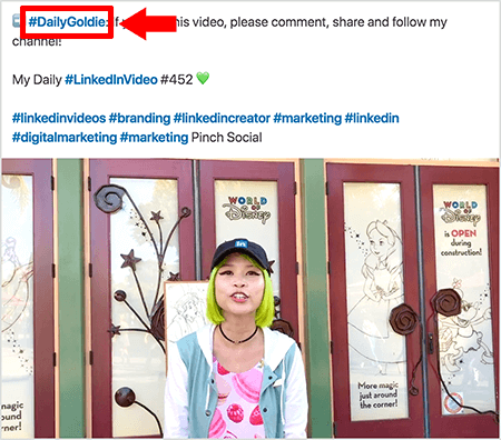 Esta es una captura de pantalla que ilustra cómo Goldie Chan usa hashtags en el texto de sus publicaciones de video de LinkedIn. Las llamadas rojas apuntan al hashtag #DailyGoldie en el texto, que es exclusivo de sus publicaciones de video y la ayuda a rastrear las acciones. La publicación también incluye otros hashtags relevantes que ayudan a las personas a encontrar su video, incluido #LinkedInVideo. En la imagen del video, Goldie se para frente a algunas puertas en una exhibición de World of Disney. Ella es una mujer asiática con cabello verde. Lleva una gorra negra de LinkedIn, una gargantilla negra, una camisa rosa con estampado de macaron y una chaqueta azul y blanca.