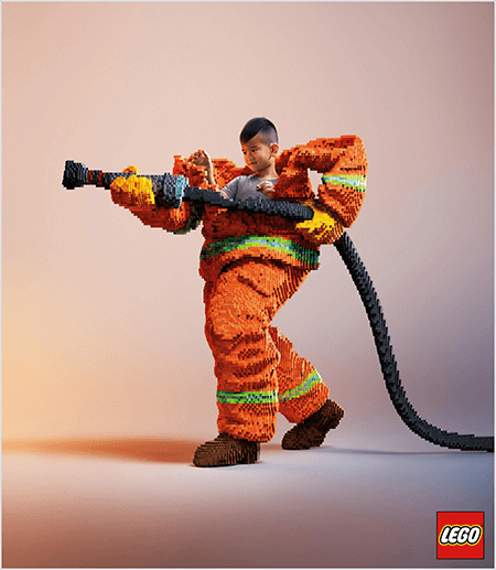 Esta es una foto de un anuncio de LEGO que muestra a un joven asiático dentro de un uniforme de bombero hecho de LEGO. El uniforme es naranja con una raya verde neón alrededor de los puños del abrigo y los pantalones. El bombero está parado con un pie hacia atrás y sosteniendo una manguera de fuego, también hecha de legos. La cabeza del niño aparece fuera de la parte superior del uniforme, que es mucho más grande que él y se detiene alrededor de los hombros. La foto fue tomada sobre un fondo neutro simple. El logotipo de LEGO aparece en un cuadro rojo en la parte inferior derecha. Talia Wolf dice que LEGO es un gran ejemplo de una marca que usa la emoción en la publicidad.
